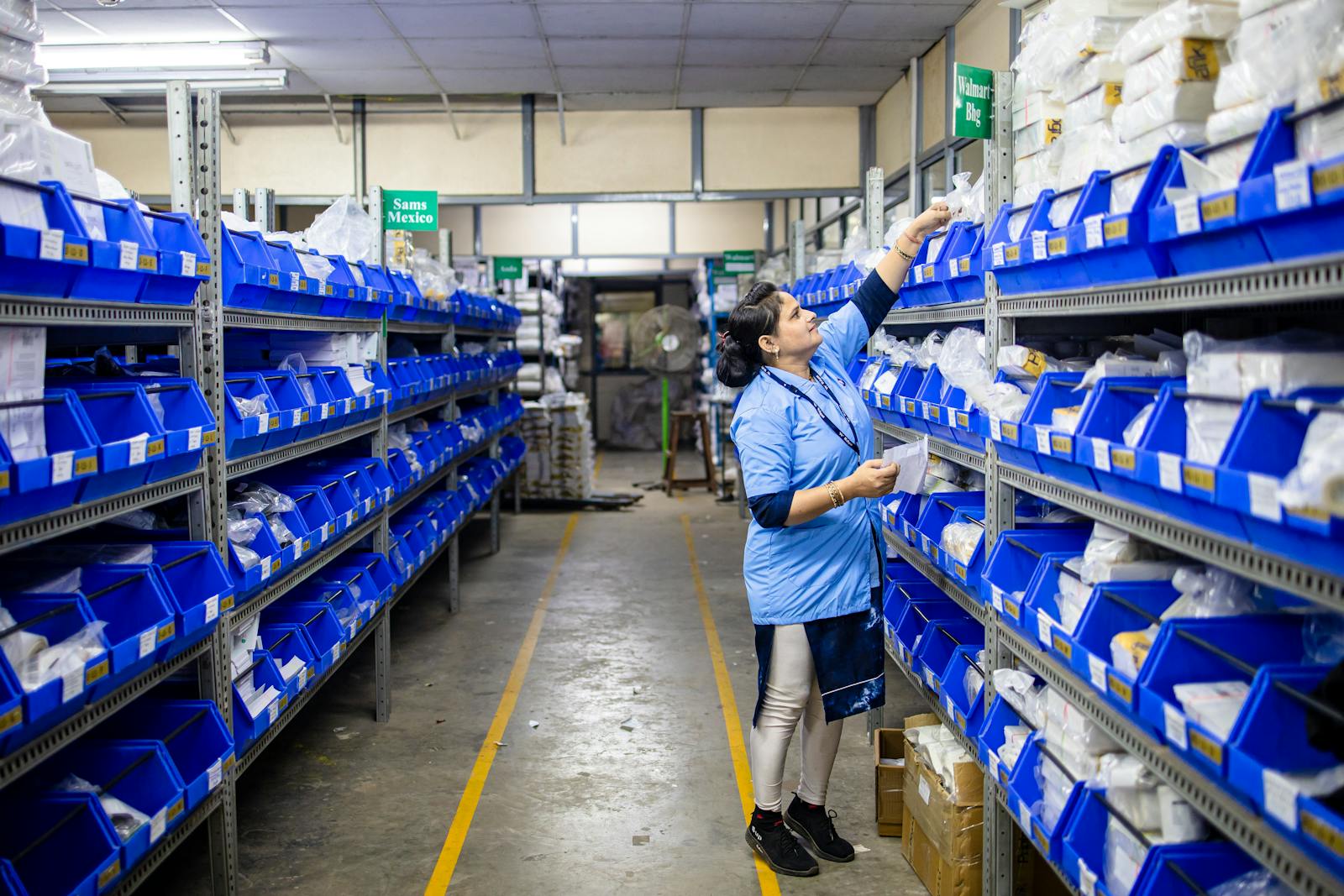 Operaria con uniforme azul revisando cajas azules en pasillo de almacén de distribución farmacéutica GDP